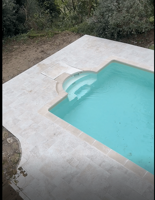 Piscine avec dallage en travertin clair, margelles assorties et escalier intérieur dans un jardin verdoyant.